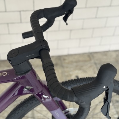 Parte frontal de uma bicicleta com guiador preto, quadro roxo e pneumáticos para terrenos irregulares.