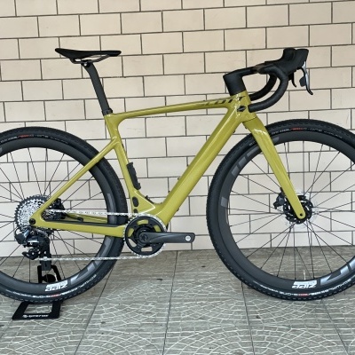 Bicicleta de estrada verde-oliva com componentes pretos e pneus volumosos junto a parede de azulejos.