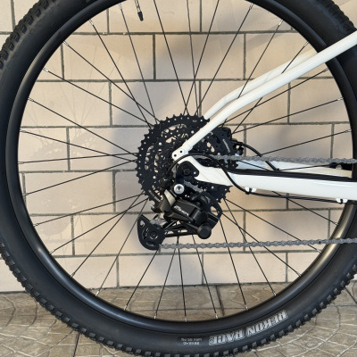 Roda traseira de bicicleta preta com pneu Maxxis e quadro branco sobre piso cerâmico e parede de azulejo.