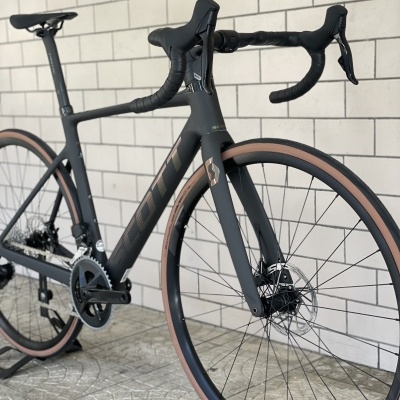 Bicicleta de estrada preta com guiador drop e pneus com faixa castanha clara encostada a parede de tijolos brancos