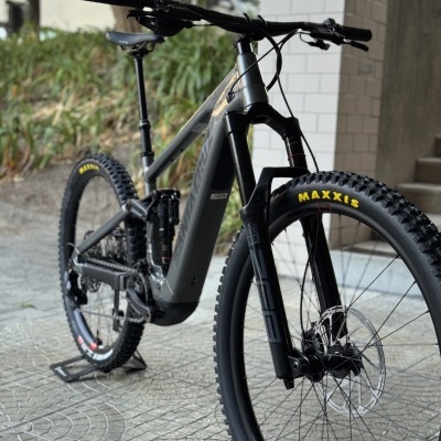 Bicicleta de montanha elétrica cinza e preta com pneus Maxxis, numa calçada ao lado de um edifício.