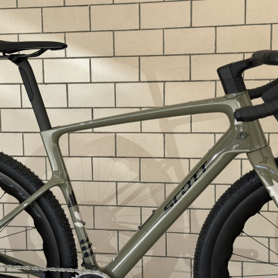 Bicicleta verde mate de corrida com selim e guiador pretos, pneus Schwalbe, encostada em azulejos bege.