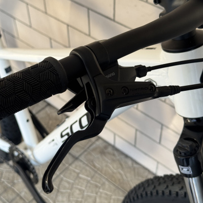 Punho preto e manete de travão TEKTRO em bicicleta branca SCOTT junto a parede de azulejos brancos.