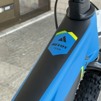 Quadro de bicicleta azul e preto com decalque REVOX TEAM em escudo azul, pneus largos e fundo com pavimento e porta de vidro