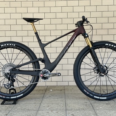 Bicicleta de montanha carbono preta com pneus Maxxis e detalhes dourados numa parede branca