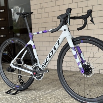 Bicicleta branca e lilás SCOTT de estrada com detalhes pretos encostada a uma parede de tijolos para venda