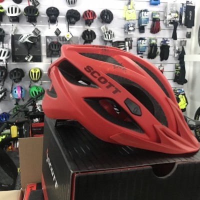 Capacete de ciclismo vermelho e preto SCOTT sobre caixa preta numa loja com vários capacetes e bicicletas