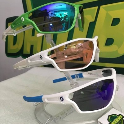 Três óculos de sol desportivos em exposição com cores verde, branco e azul, cada um com lentes coloridas distintas.