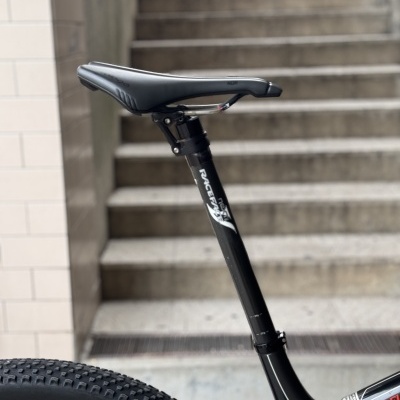 Assento de bicicleta preto montado em quadro de carbono preto com detalhes vermelhos e branco, roda traseira com pneu de cravos e escadas ao fundo