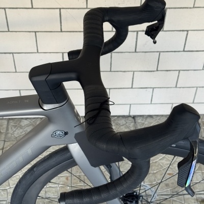 Guiador de bicicleta preto com fita aderente e quadro cinza numa parede branca e chão com azulejos industriais