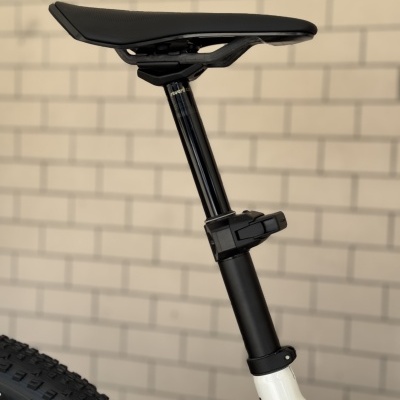 Selim de bicicleta preto com garfo ajustável e quadro branco sobre fundo de parede de tijolos.