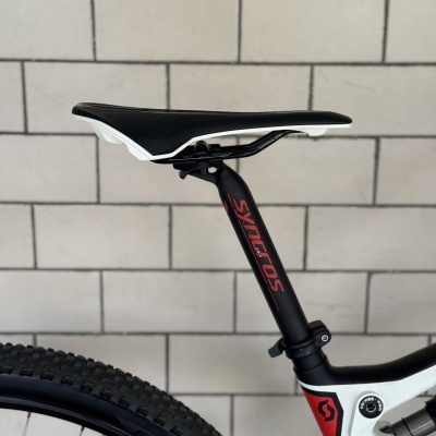 Banco de bicicleta preto e branco com canote preto com texto vermelho e pneus de borracha preta junto a parede de azulejo branco