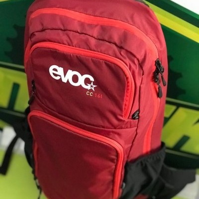 Mochila vermelha evoc CC 16L com múltiplos bolsos e alças pretas, sobre fundo verde amarelo preto.
