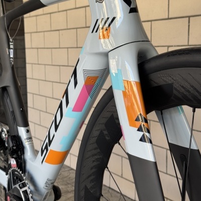 Quadro de bicicleta branca com padrões coloridos e roda preta junto a parede de azulejos bege