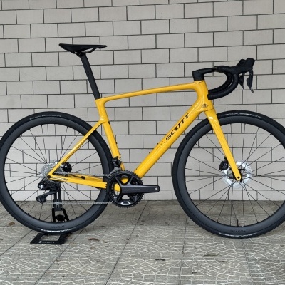 Bicicleta de estrada amarela SCOTT com rodas pretas e selim preto encostada numa parede de blocos cinza.