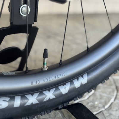 Roda de bicicleta com aro cinza, pneu preto e suporte preto sobre chão de pedra