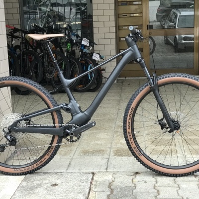 Bicicleta de montanha elétrica cinza com pneus castanhos no pavimento de pedra em frente a um prédio