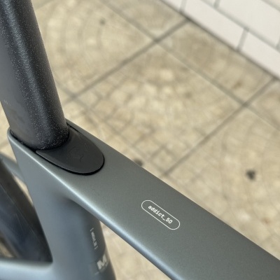Detalhe de bicicleta cinzenta com texto addict_50 sobre chão e parede de azulejos.