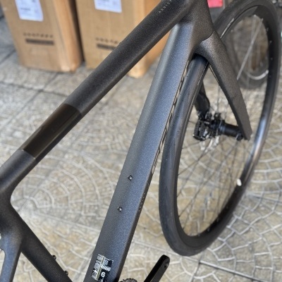 Bicicleta de carbono cinzenta com roda traseira preta sobre pavimento com caixas ao fundo