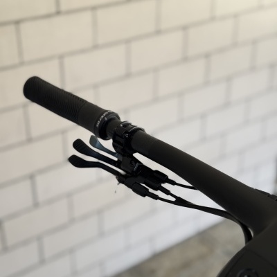 Punho esquerdo de guiador de bicicleta preto com manetas e cabos, parede de tijolos ao fundo