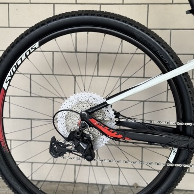Roda traseira de bicicleta de montanha com pneu preto e aro SYNCROS, cassete metálico, desviador SRAM, quadro branco e preto com detalhes vermelhos, parede de azulejos brancos