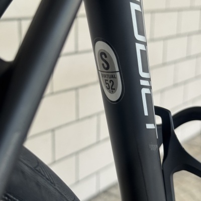 Quadro de bicicleta preto fosco com autocolante de tamanho e parede branca azulejada ao fundo.