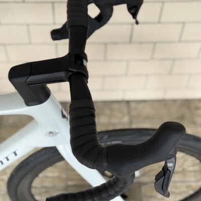 Guiador de bicicleta de estrada preto com fita texturada e manetes integradas, quadro branco, roda preta, parede de azulejos claros ao fundo