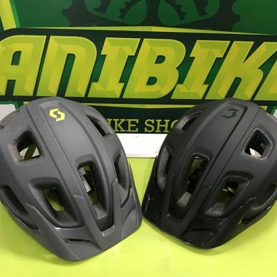 Dois capacetes de ciclismo SCOTT, um cinza e um preto, com ventilação, sobre superfície amarela na loja DANIBIKES