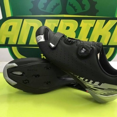 Ténis de ciclismo pretos com detalhe branco e sola preta exibidos sobre superfície amarela junto a fundo com texto