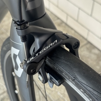 Travão dianteiro negro de bicicleta com texto ULTEGRA numa roda com pneu preto e aro cinzento, na estrutura cinzento escuro com padrão geométrico.