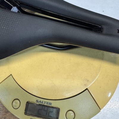 Sela preta de bicicleta sobre balança digital amarela mostrando 242 g.