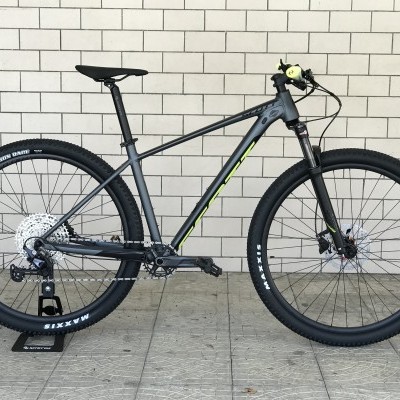 Bicicleta de montanha cinzenta escura com detalhes amarelos e pneus Maxxis contra parede branca