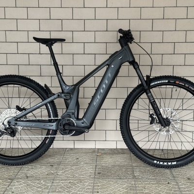 Bicicleta de montanha elétrica cinza SCOTT encostada numa parede de tijolos bege
