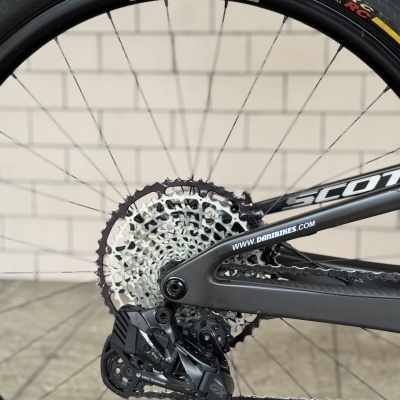 Pneu de bicicleta preto com aro e sistema de mudanças visíveis, texto em amarelo, branco e preto, quadro cinza, fundo de parede clara