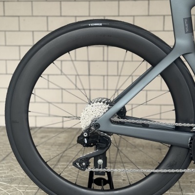 Roda traseira de bicicleta com pneu preto e quadro cinza azulado junto a chão de azulejos e parede de tijolos bege