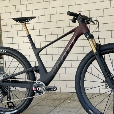 Bicicleta de montanha carbono preta com detalhes dourados e pneus Maxxis encostada a parede