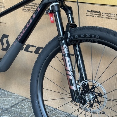 Parte da frente de uma bicicleta de montanha preta com suspensão e pneu Maxxis, com caixa de cartão ao fundo.
