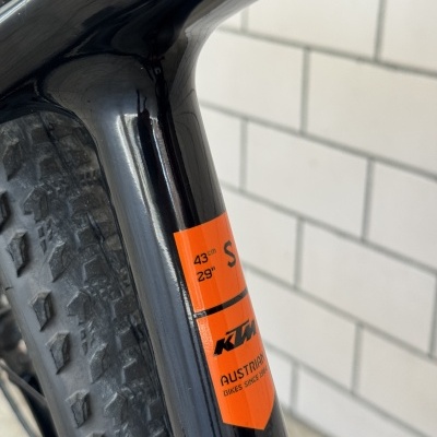 Quadro de bicicleta preto com autocolante laranja e pneu com rasto volumoso em fundo de azulejos brancos.