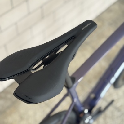 Assento de bicicleta preto com textos AMANCH e PARTICLE FLOW, bicicleta lilás em ambiente interior
