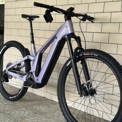 Bicicleta elétrica de montanha lilás com pneus Maxxis em parede de azulejos claros