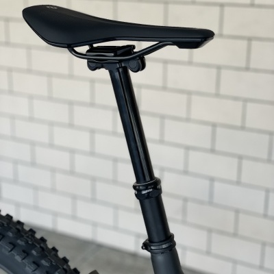 Selim preto de bicicleta com cana e pneu, parede de tijolos brancos ao fundo