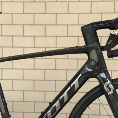 Bicicleta de estrada preta em carbono com logo Scott e pneus Schwalbe junto a parede de azulejos.