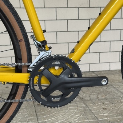 Quadro de bicicleta amarelo com sistema de pedais e pneu castanho e preto junto a parede de tijolos.