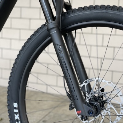 Roda dianteira de bicicleta com suspensão e pneu preto MAXXIS junto a parede de tijolos