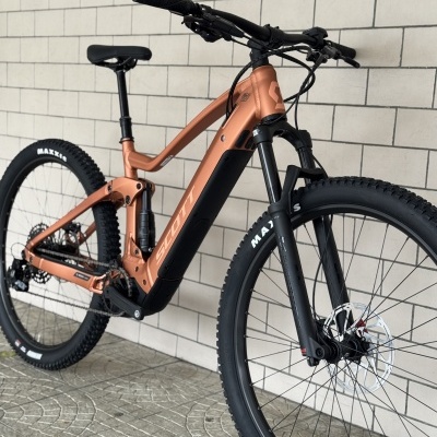 Bicicleta de montanha elétrica bronze com pneus Maxxis e suspensão, encostada numa parede branca de tijolos.