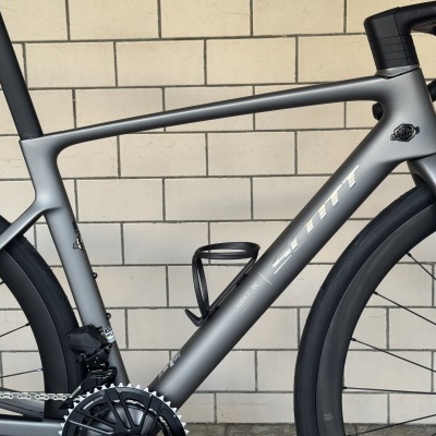 Bicicleta de estrada Scott cinzenta com texto ADDICT RC e DANTBIKES contra parede de azulejos claros