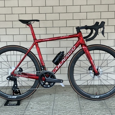 Bicicleta de estrada vermelha metalizada com componentes pretos apoiada em suporte diante de parede branca azulejada