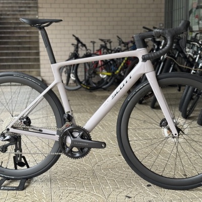 Bicicleta de estrada cinza claro com rodas pretas e selim preto num espaço interior com outras bicicletas