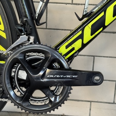 Parte do quadro de uma bicicleta de estrada preta e amarela com pedivela e desviador DURA-ACE, em chão de tijoleira e pedra.