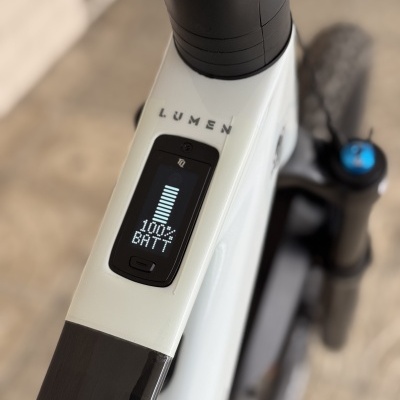 Painel digital a mostrar bateria a 100% numa bicicleta elétrica branca e preta com texto LUMEN no quadro.